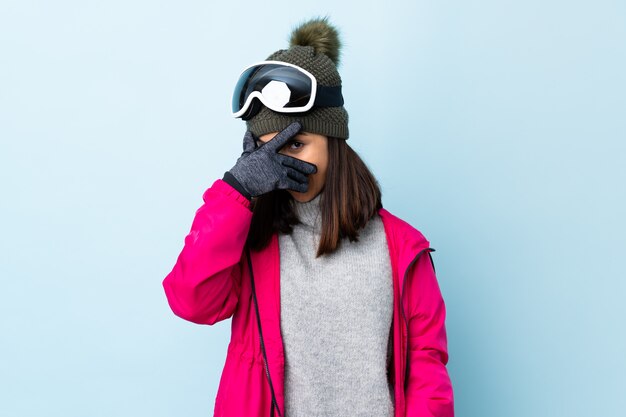 La ragazza dello sciatore della corsa mista con i vetri di snowboard sopra il rivestimento blu della parete osserva a mano e sorridendo.