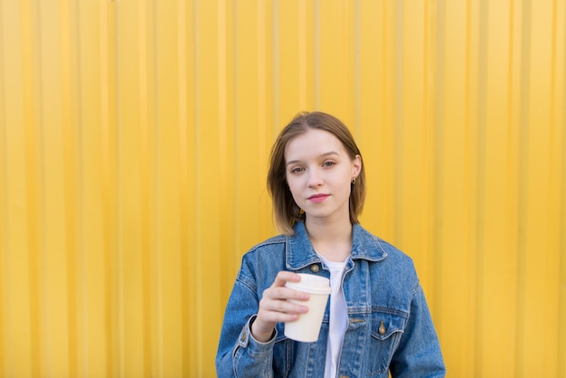 La ragazza dello studente sta con la tazza di caffè in sue mani su fondo giallo ed esamina la macchina fotografica.