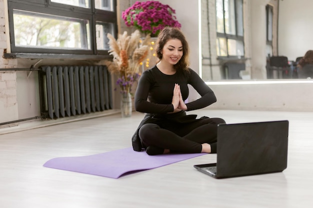 La ragazza dello yoga guarda il tutorial sullo sport del laptop Donna sportiva sorridente in forma in abiti sportivi in camera guardando video di yoga sul laptop mentre si è seduti su un tappetino fitness