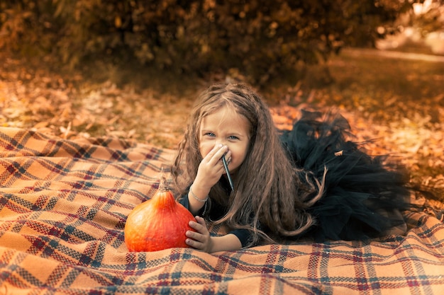 La ragazza di Halloween con i capelli lunghi che indossa un vestito nero si siede su una coperta arancione nel parco e disegna un