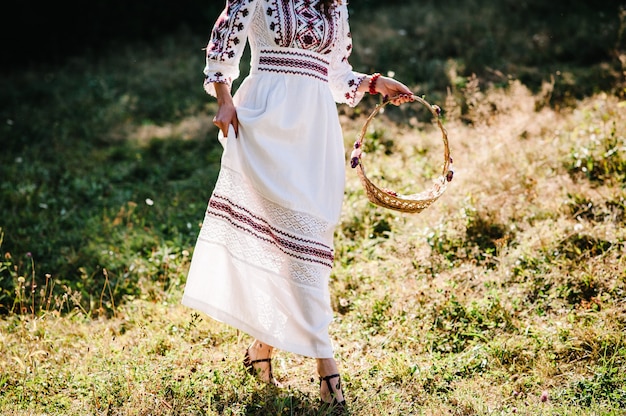 La ragazza di vista dal basso con fiori nel cestino a piedi nel parco, preparazione della damigella d'onore al matrimonio in giardino, cortile, all'aperto. Stile rustico ucraino: sposa in abito ricamato sulla natura.