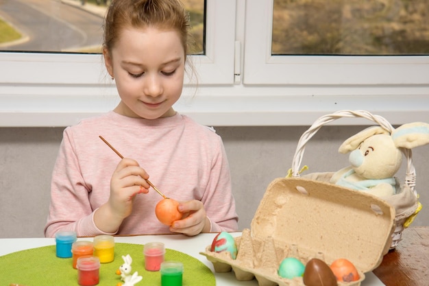 La ragazza dipinge le uova al tavolo vicino alla finestra per Pasqua con un coniglietto di Pasqua