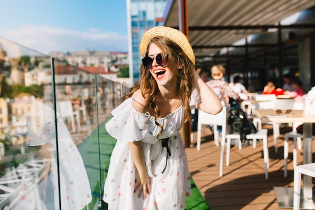 La ragazza divertente con capelli lunghi in occhiali da sole sta sulla terrazza in caffè. Indossa un abito bianco con spalle scoperte, rossetto rosso e cappello. Lei è in posa per la fotocamera.