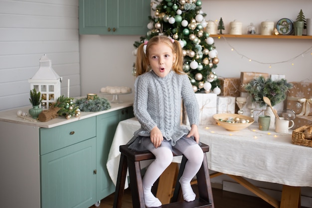 La ragazza divertente sta sedendosi nella cucina vicino all'albero di Natale decorato all'interno.