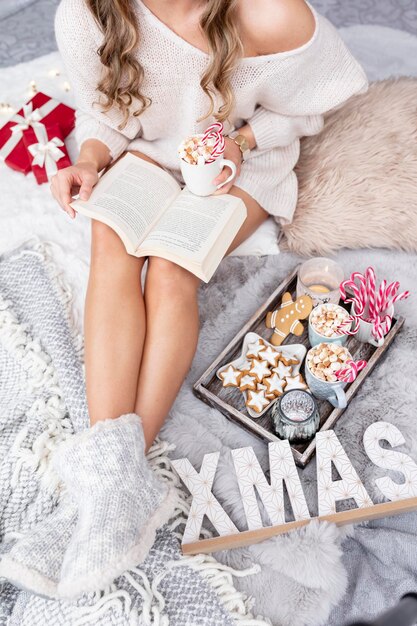 La ragazza è seduta in un'atmosfera natalizia, beve una bevanda calda e legge un libro.