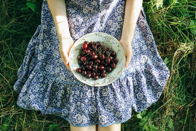 La ragazza è seduta sull'erba in abito vintage blu. La donna sta tenendo le ciliegie. Piatto di frutta estiva rustica. Concetto di stile di vita sano cibo ecologico vegetariano.