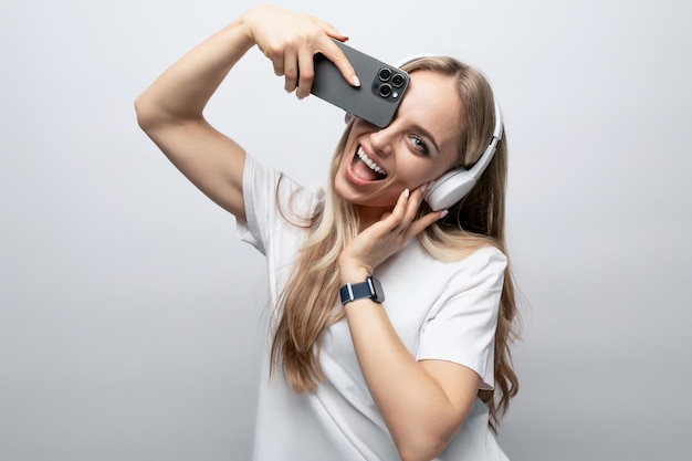 La ragazza fa smorfie in cuffie wireless con uno smartphone in mano su uno sfondo bianco