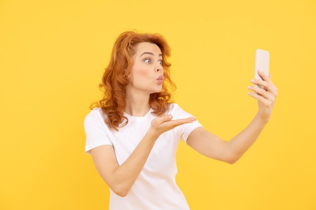 La ragazza fa un gesto di bacio della mano al dispositivo mobile con sfondo giallo del telefono con fotocamera