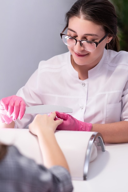 La ragazza fa una manicure in un salone di bellezza Cura delle unghie Una manicure in guanti rosa archivia le unghie di un cliente Procedure cosmetiche