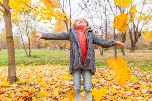 La ragazza felice che gioca sotto le foglie gialle che cadono nel bellissimo parco autunnale sulla natura cammina all'aperto. Il bambino vomita foglie d'acero arancioni autunnali.