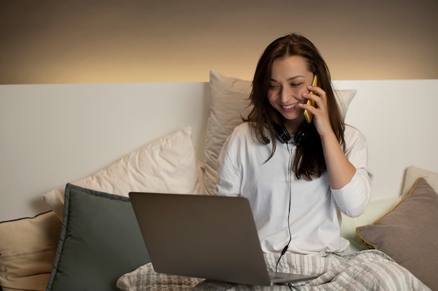 La ragazza felice che si siede nel letto, sorridendo, lavora al computer portatile da casa, parlando al telefono