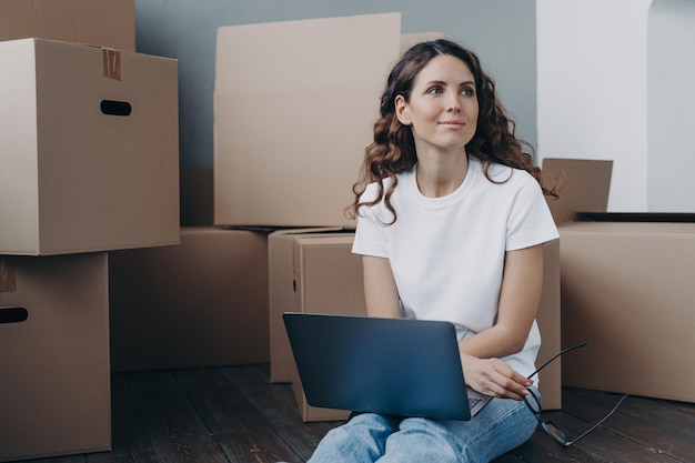 La ragazza felice con il computer portatile si siede sul pavimento vicino alle scatole di cartone sognando il nuovo concetto di rilocazione della casa