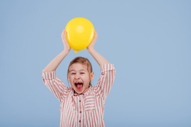 La ragazza felice del bambino sostiene il pallone giallo vestito in camicia a strisce isolata su fondo blu copia s...