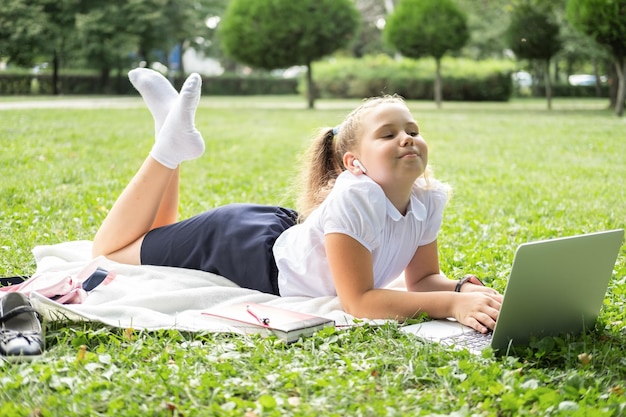 la ragazza felice del bambino sull'uniforme scolastica con le cuffie wireless ha una lezione online sul laptop sul prato