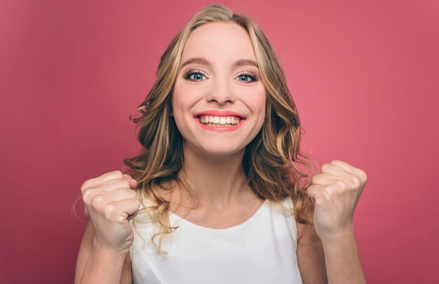 La ragazza felice e attraente sta guardando dritto e tiene i suoi pugni. Anche lei sta sorridendo. La giovane donna è molto allegra. Isolato su muro rosa