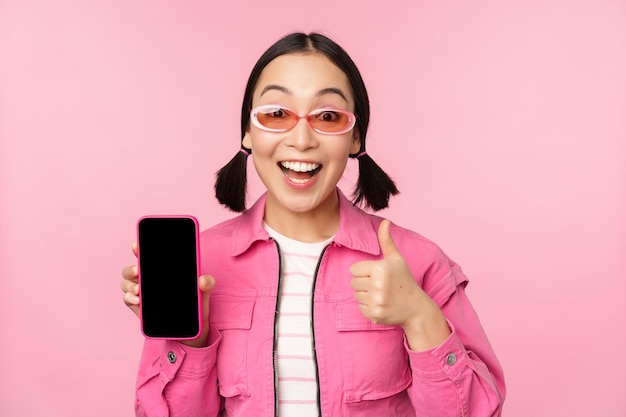La ragazza felice ed elegante consiglia l'applicazione sul telefono cellulare Modello femminile asiatico sorridente che mostra lo schermo dello smartphone e il pollice in su in piedi su sfondo rosa