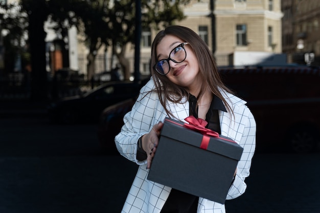 La ragazza felice imbarazzata con il regalo nelle sue mani sorride. La giovane donna sta tenendo ordinatamente la scatola nera con l'arco rosso.