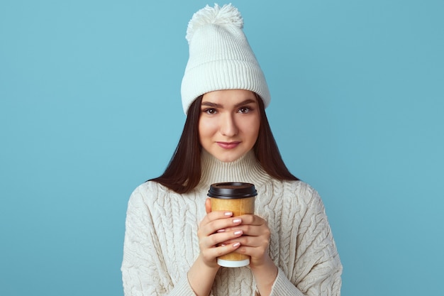 La ragazza felice in cappello bianco e maglione caldo bianco tiene una tazza di caffè da asporto