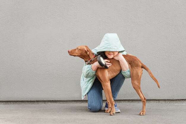 La ragazza felice in una giacca abbraccia un bellissimo cane marrone su un grigio