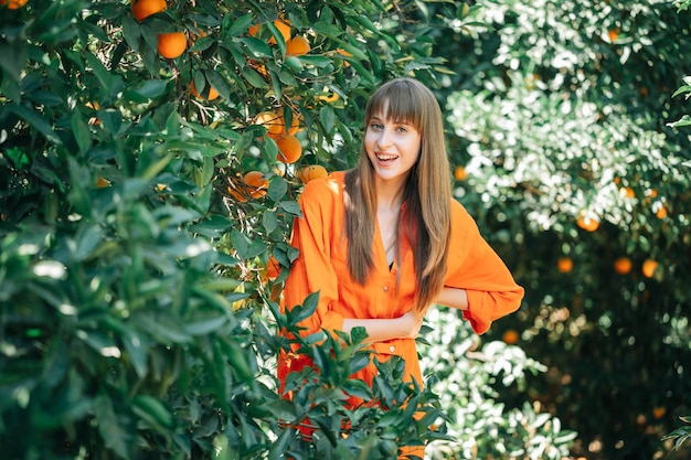 La ragazza felice in vestito arancione sta posando alla macchina fotografica unendo le mani sulla vita nel giardino d'arancio