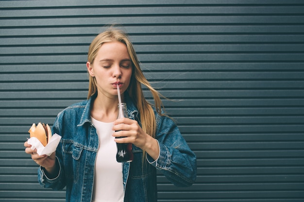 La ragazza felice mangia gli alimenti a rapida preparazione e beve la cola vicino vicino alla parete grigia