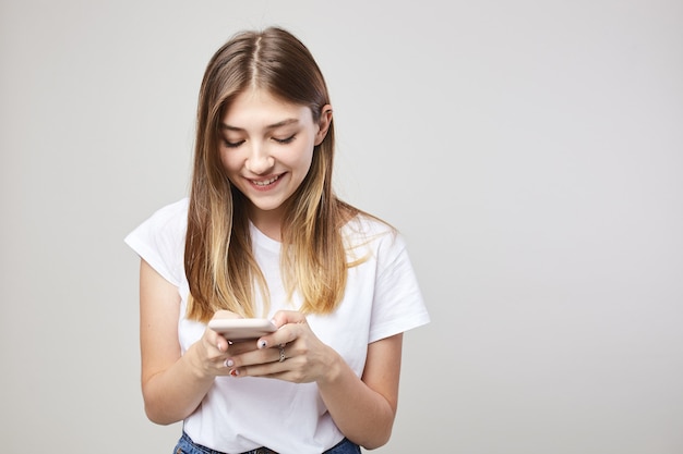 La ragazza felice sorride e usa il telefono cellulare su uno sfondo bianco in studio