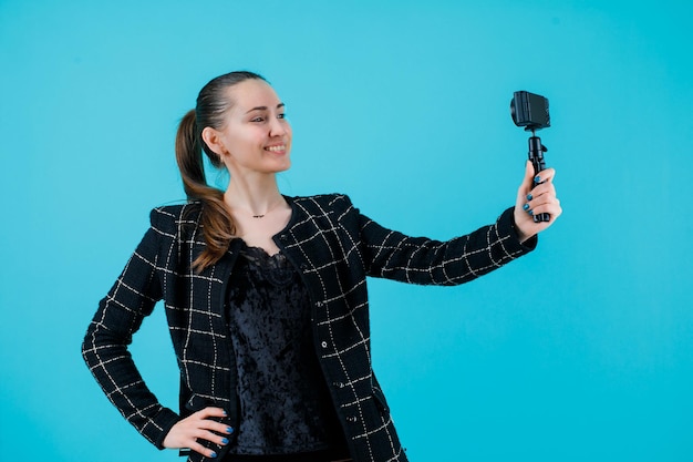 La ragazza felice sta prendendo selfie con la mini macchina fotografica mettendo la mano sulla vita su fondo blu