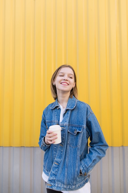 La ragazza felice sta stando su fondo giallo e sta bevendo il caffè.