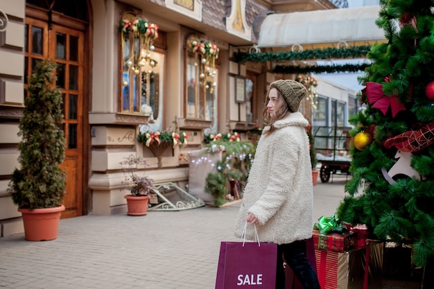 La ragazza felice tiene i sacchetti di carta con il simbolo della vendita nei negozi con le vendite a Natale in giro per la città