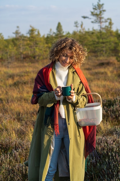 La ragazza gioiosa in trincea mano calda su una tazza di tè calda trascorre il fine settimana godendosi l'autunno soleggiato in campagna