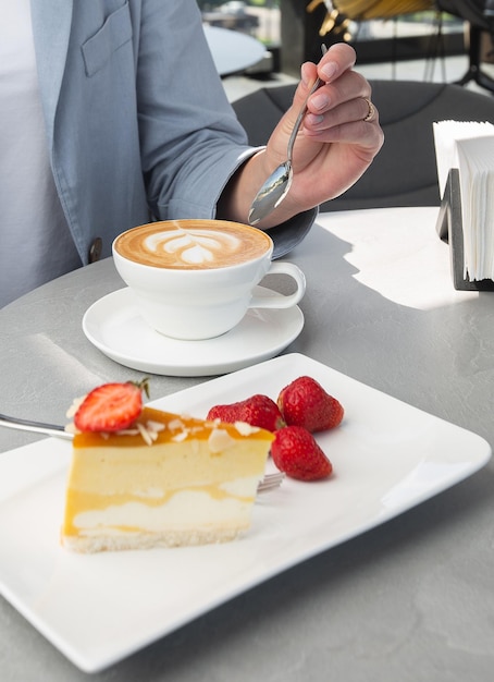 La ragazza gode della classica cheesecake con fragole fresche con una tazza di caffè sul tavolo bianco. Vista ravvicinata. Colazione gustosa. Pezzo di torta sul piatto, tazza bianca su fondo di marmo bianco.