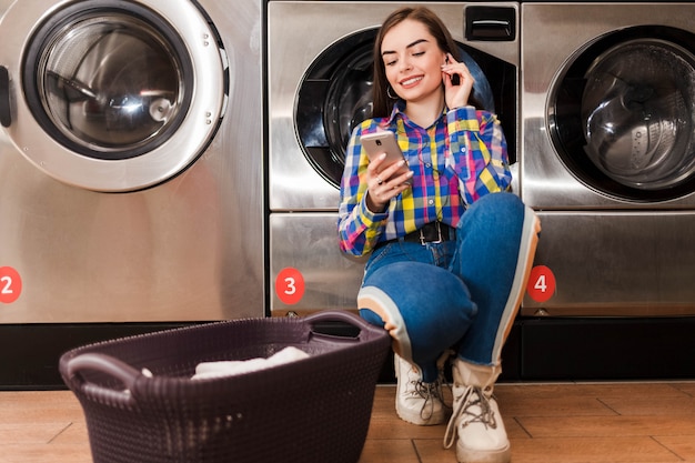 La ragazza graziosa ascolta la musica che si appoggia su una lavatrice nella lavanderia pubblica