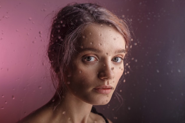 la ragazza graziosa esamina la macchina fotografica su priorità bassa viola. gocce d&#39;acqua sfocate corrono giù per il vetro davanti al suo viso