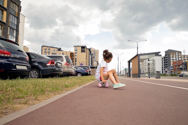 La ragazza guida uno skateboard su un marciapiede della città vicino a un parcheggio e ad edifici.