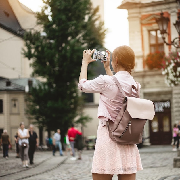 La ragazza hipster con lo zaino tiene lo smartphone Il viaggiatore fotografa il paesaggio urbano al tramonto Stile di vita