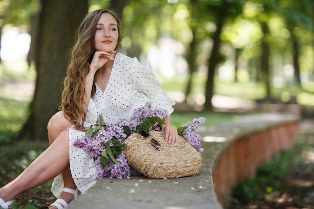 La ragazza in abito bianco e occhiali da sole tiene tra le mani un cesto di vimini con fiori. Cesto con lillà. Ragazza e fiori. Siediti con un cesto di lillà nelle mani. floristica