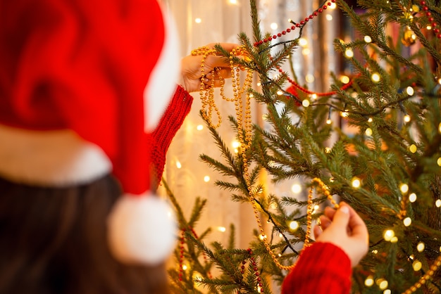 La ragazza in cappello di natale rosso e bianco brillante decora l'albero di abete con perline