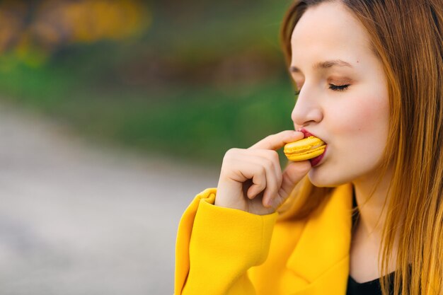 La ragazza in cappotto giallo mangia il maccherone giallo. trucco. avvicinamento.