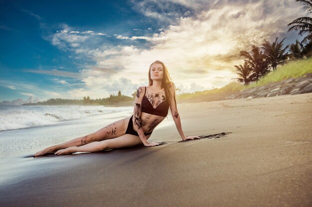 La ragazza in costume da bagno ricoperta di sabbia nera è sdraiata sulla spiaggia al tramonto e nell'oceano