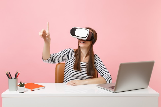 La ragazza in cuffia della realtà virtuale sulla testa tocca qualcosa come premere il pulsante o indicando lo schermo virtuale mobile lavora alla scrivania con il computer portatile isolato su sfondo rosa. Carriera di affari di successo.