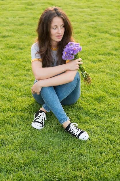 La ragazza in jeans si siede sul prato verde che gode del mazzo dei fiori freschi, concetto di vacanza estiva.