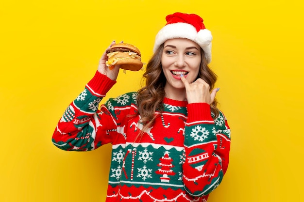 la ragazza in maglione di Natale e cappello di Babbo Natale tiene un grande cheeseburger e lecca la salsa