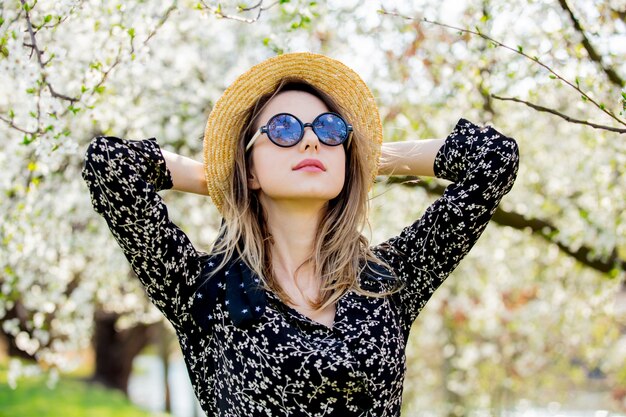 La ragazza in occhiali da sole e cappello sta vicino ad un albero di fioritura