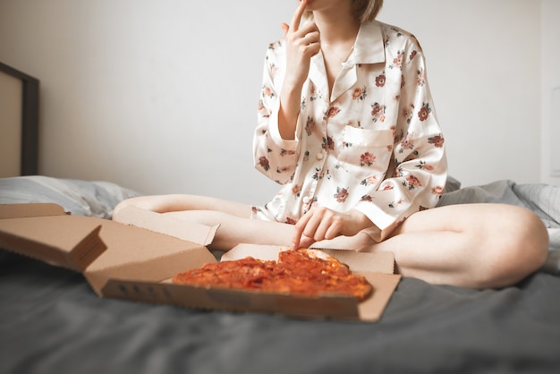 La ragazza in pigiama si siede sul letto in camera da letto e mangia una pizza dalla scatola.