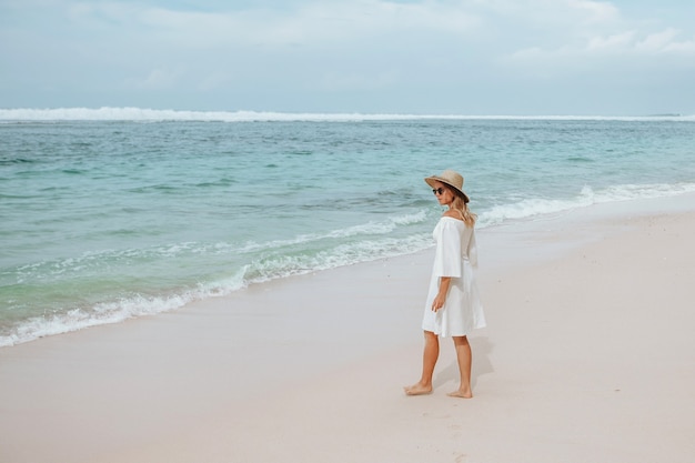 La ragazza in un vestito bianco cammina su una spiaggia bianca in un cappello e occhiali da sole