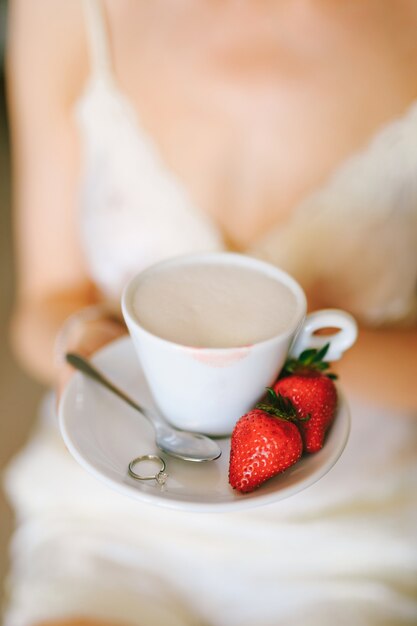 La ragazza in un vestito bianco tiene nelle sue mani un piattino con una tazza di cappuccino due fragole a