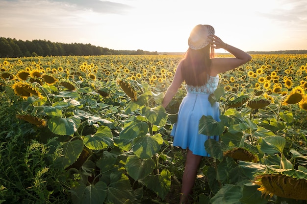 La ragazza in un vestito blu lascia con il cappello nel campo dei girasoli sul tramonto Follow me concept