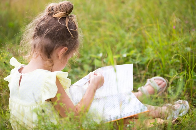 La ragazza in un vestito giallo si siede nell'erba su una coperta in un campo e legge un libro di carta. Giornata internazionale dei bambini. L'ora legale, l'infanzia, l'istruzione e l'intrattenimento, il nucleo del cottage. Copia spazio
