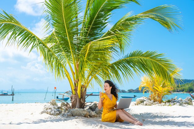 La ragazza in un vestito giallo su una spiaggia sabbiosa tropicale lavora ad un computer portatile e beve il mango fresco. Lavoro a distanza, libero professionista. Funziona in vacanza.