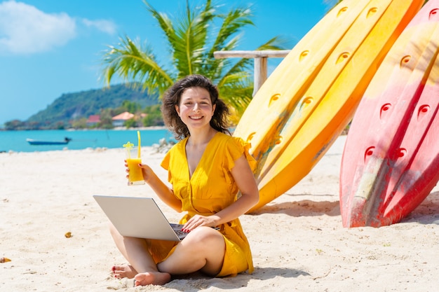 La ragazza in un vestito giallo su una spiaggia sabbiosa tropicale lavora ad un computer portatile e beve il mango fresco. Lavoro a distanza, libero professionista. Funziona in vacanza.